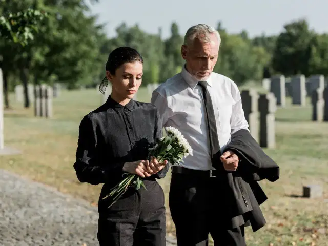 Para idąca przez cmentarz, kobieta w czarnej sukience trzyma bukiet białych kwiatów, mężczyzna w białej koszuli i czarnym krawacie niesie marynarkę.