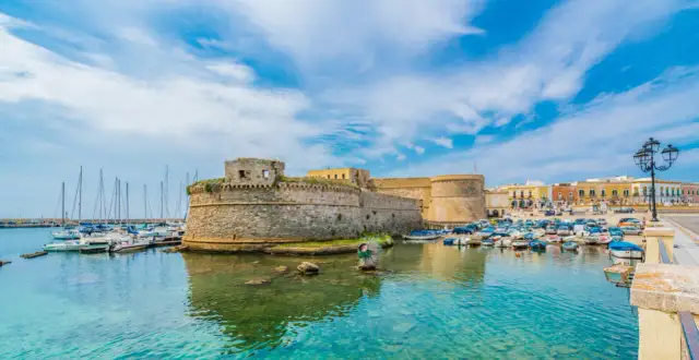 A stone castle overlooks a harbor filled with boats and yachts under a bright blue sky.