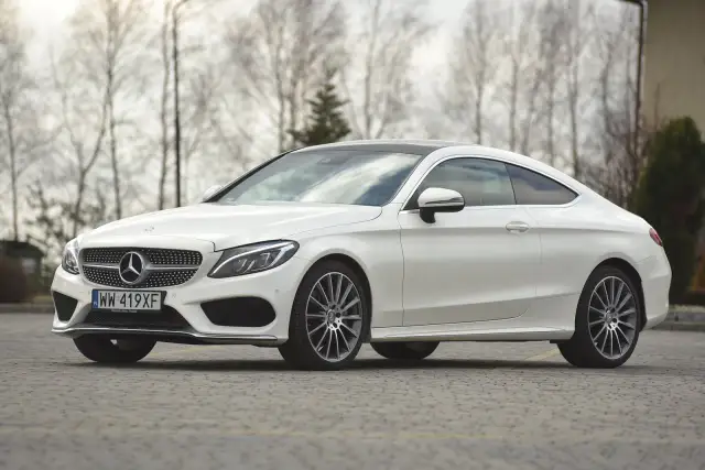 Biały Mercedes C klasa coupe z czarnym dachem, zaparkowany na brukowanej nawierzchni, z drzewami w tle.