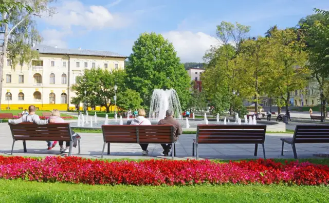 Park z fontanną i ludźmi siedzącymi na ławkach, w tle budynek.