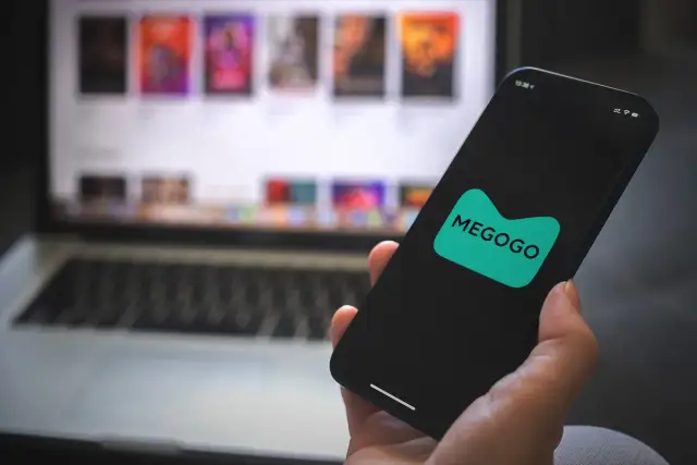 Dłoń trzyma smartfon z logo MEGOGO. Na ekranie laptopa w tle widać okładki filmów.