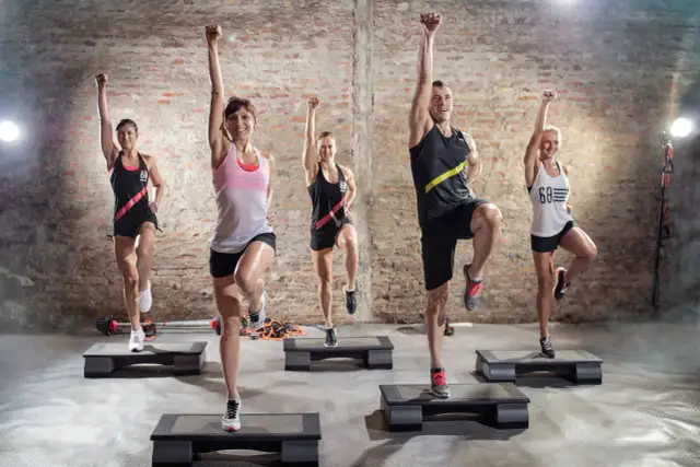 Grupa osób ćwiczy na stepach, podnosząc kolana i ręce. To świetny przykład, co to jest trening aerobowy.