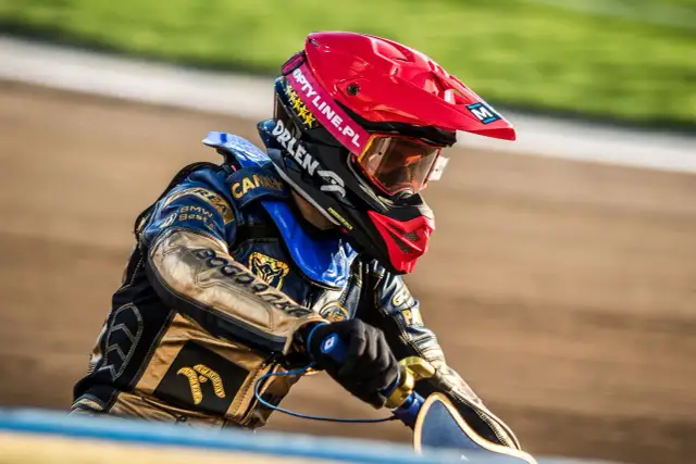 Żużlowiec w kasku i kombinezonie czeka na start. Jego strój zdobią logotypy sponsorów, a na torze liczą się pge ekstraliga statystyki.