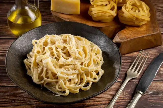 Kremowe fettuccine alfredo na czarnym talerzu, obok surowy makaron, ser i oliwa.