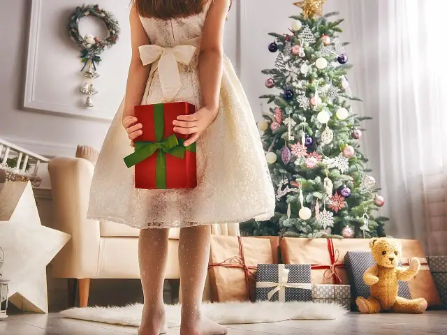 Dziewczynka w białej sukience trzyma czerwony prezent przy choince.