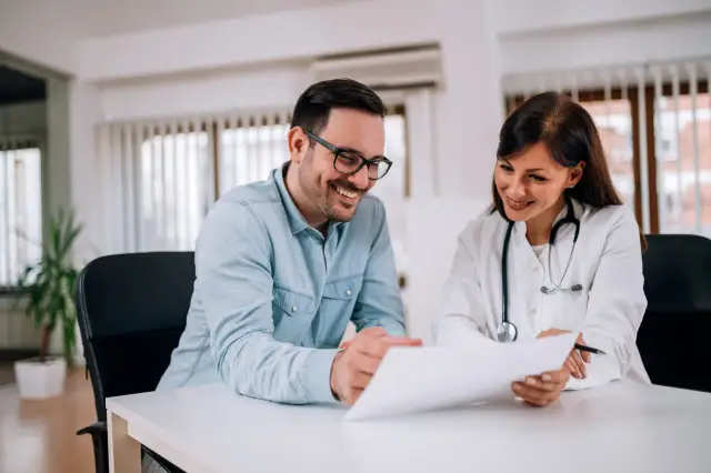 Lekarka w białym fartuchu i pacjent w okularach omawiają dokumenty przy biurku.