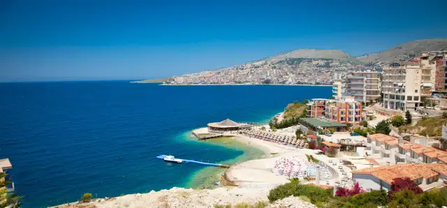 Widok na wybrzeże Sarandy, z turkusową wodą, plażą z parasolami i budynkami. W oddali widać panoramę miasta, przypominającą widok z Tirany.
