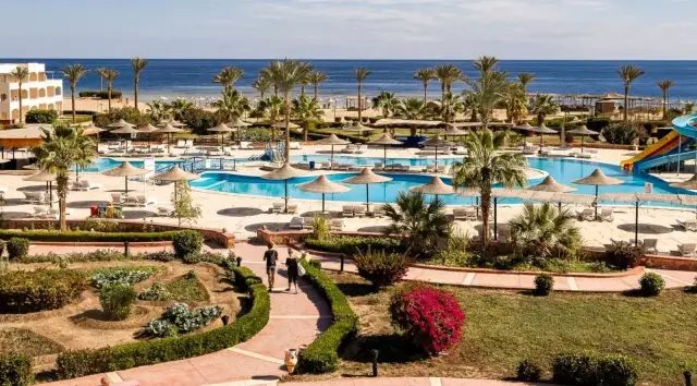 Widok na basen i plażę w kurorcie z palmami i leżakami.