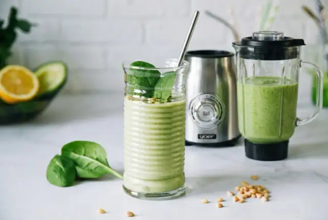Zielony koktajl owocowo-warzywny w szklance z metalową słomką, obok blendera i składników.