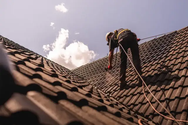 Pracownik w kasku i uprzęży zabezpieczającej wykonuje renowację dachu, natryskując dachówkę.