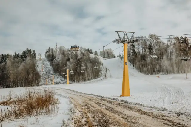 Wyciąg krzesełkowy na ośnieżonym stoku narciarskim z widocznym budynkiem na wzgórzu.