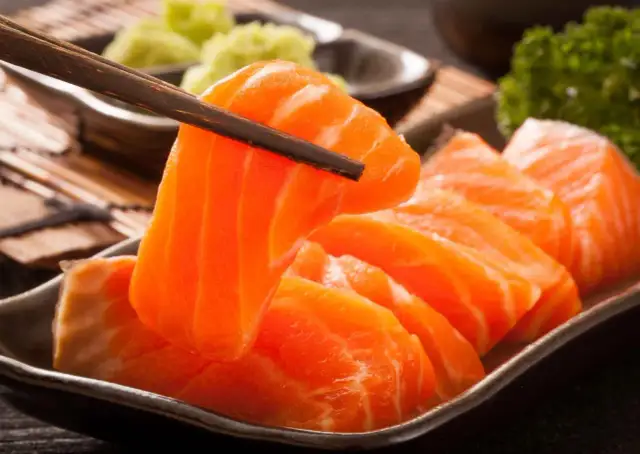 Sashimi co to? Świeże, pomarańczowe kawałki łososia podniesione pałeczkami, obok zielonej pasty wasabi i natki pietruszki.