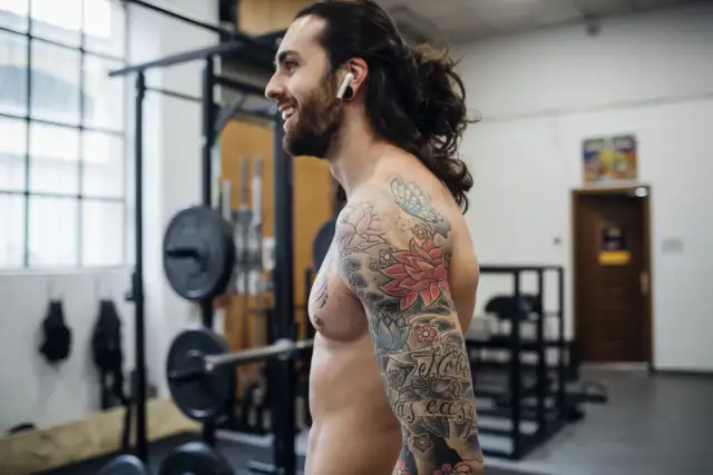 Uśmiechnięty mężczyzna z tatuażem na ramieniu w siłowni. Zastanawia się, ile dni po zrobieniu tatuażu można ćwiczyć.