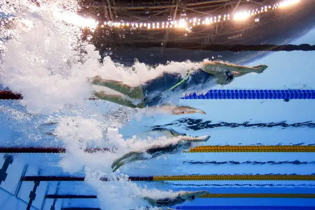 Pływacy w stylu dowolnym ścigają się pod wodą, tworząc fale. Widać różne style pływania, od szybkiego kraula po bardziej techniczne style.