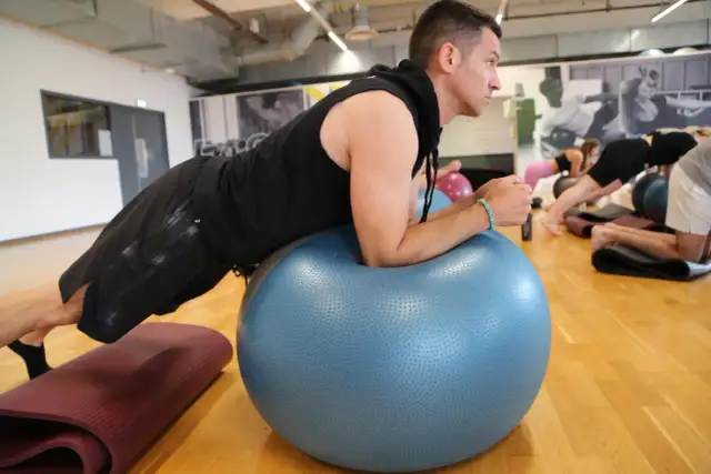 Mężczyzna w czarnej bluzie i spodenkach ćwiczy na dużej, niebieskiej piłce fitness.