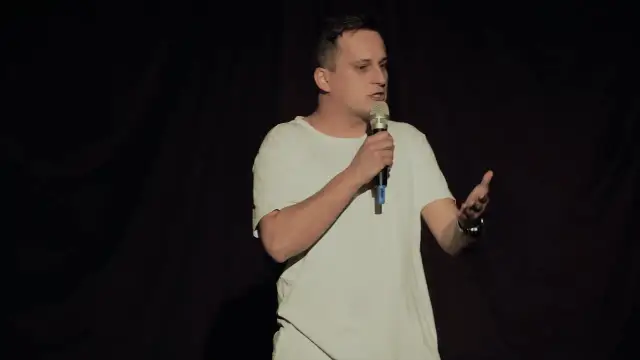 Mężczyzna w białej koszulce stoi na scenie z mikrofonem w dłoni, gestykulując.