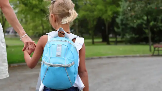 Dziecko w niebieskim plecaku trzyma za rękę dorosłego, idąc przez park.