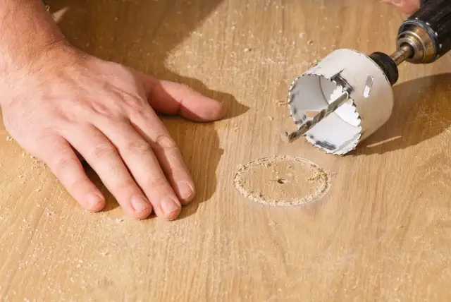 Dłoń przytrzymuje drewno podczas wiercenia otworów wiertłem koronowym.