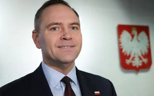 Polityk w garniturze z polskim godłem w tle.