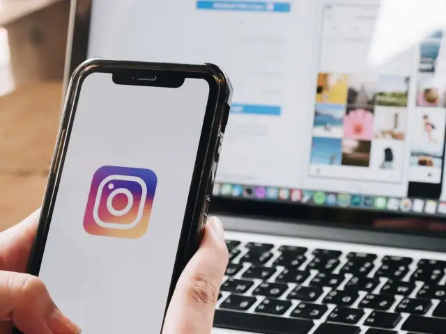 Telefon z logo Instagrama w dłoni, obok laptop z otwartą aplikacją. Dowiedz się, jak zmienić dźwięk powiadomień na Instagramie.