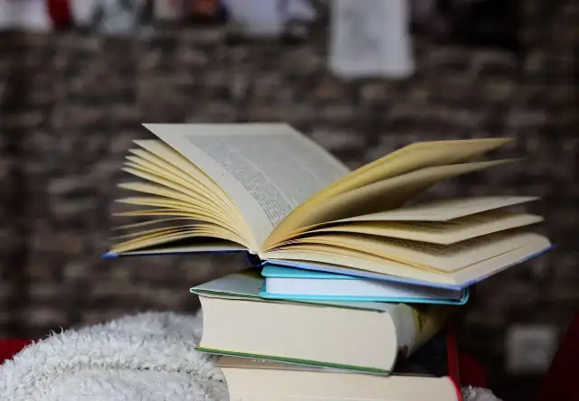 Otwarta książka leży na stosie innych książek, na tle ceglanej ściany.