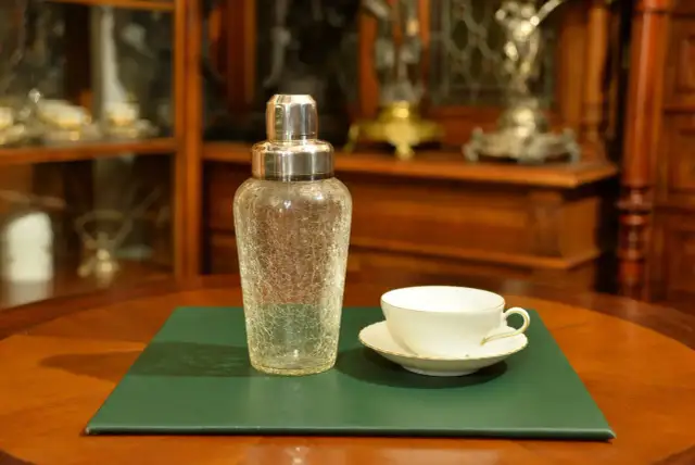 Szklany shaker do koktajli i biała filiżanka z podstawkiem na zielonej podkładce.