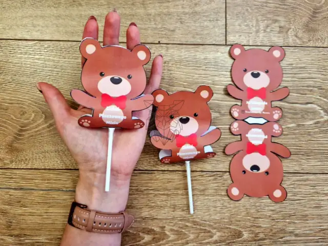 Dłoń trzymająca patyczek z papierowym misiem. Obok leżą kolejne misie.