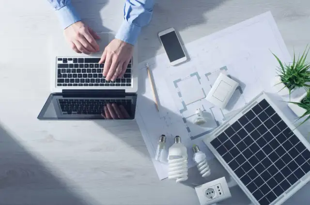 Analiza sprawności paneli fotowoltaicznych na laptopie. Na biurku projekt, żarówki, gniazdko i telefon.