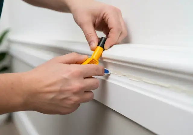 Dłonie przyklejają białą listwę do ściany za pomocą żółtego aplikatora z klejem.