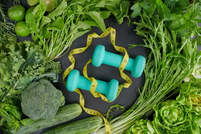 Dwie turkusowe hantle owinięte żółtą miarką, otoczone świeżymi warzywami i ziołami.