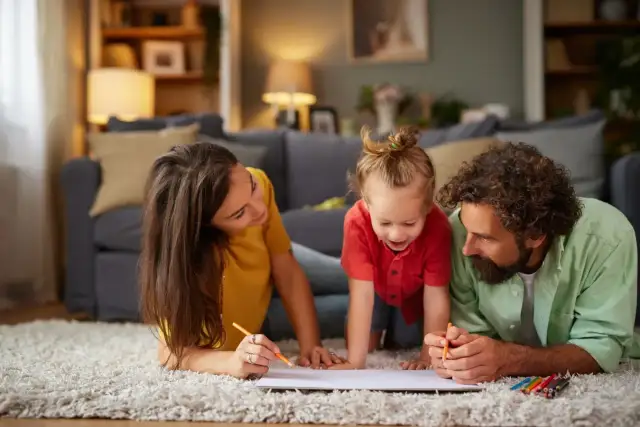 Rodzina rysuje kredkami na kartce papieru leżąc na dywanie w salonie.
