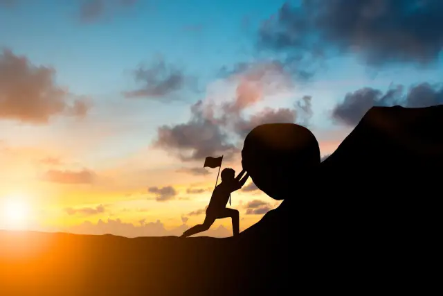 Sylwetka człowieka pchającego kamień pod górę z flagą na tle zachodzącego słońca.