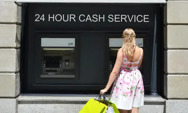 Kobieta w letniej sukience z zakupami przed bankomatem. Czy można wypłacić 20 zł z bankomatu PKO?