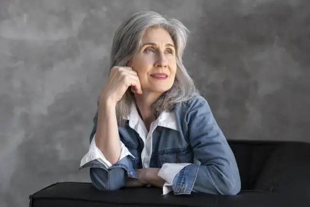 Kobieta z siwymi włosami, oparta o czarną kanapę, zamyślona. Może myśli o ziołach na pamięć, które pomagają jej zachować jasność umysłu.