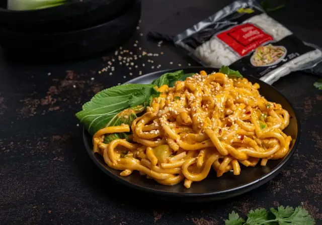 Grube, pomarańczowe kluski biang biang w sosie, posypane sezamem i chili, z zielonymi liśćmi bok choy.