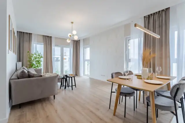 Nowoczesny salon z jadalnią. Szara kanapa, drewniany stół z krzesłami, ozdobna ściana i okna. Kto to? Flipper, bo tak nazywa się ten styl.