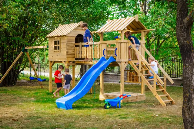 Dzieci bawią się na drewnianym placu zabaw ze zjeżdżalnią i huśtawkami w ogrodzie.