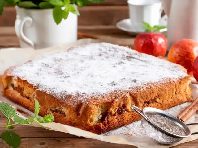 Puszysty biszkopt z jabłkami, posypany cukrem pudrem, obok jabłka i cynamon.