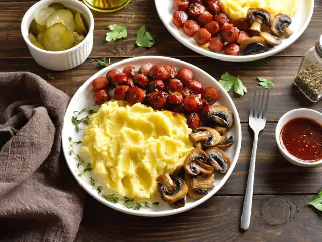 Talerz z purée ziemniaczanym, kiełbaskami i pieczarkami, obok miseczka z piklami i sosem.