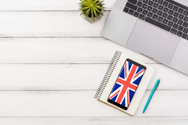 Laptop, notes, telefon z flagą Wielkiej Brytanii i długopis na białym drewnianym tle.