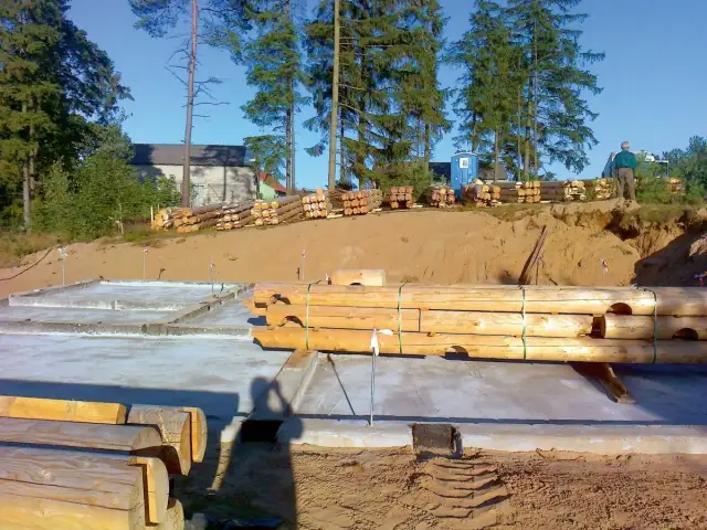 Budowa domu z bali: fundamenty, stosy drewna i pracownik na budowie.