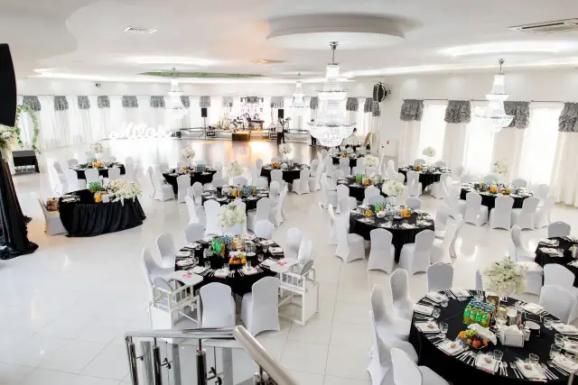 Elegancka sala weselna z czarnymi stołami, białymi krzesłami i kryształowymi żyrandolami.