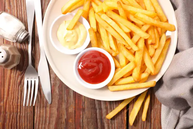 Talerz ze złotymi frytkami, ketchupem, majonezem, solniczką i sztućcami na drewnianym tle.