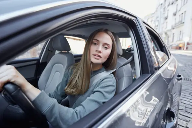 Młoda kobieta, współwłaściciel samochodu poniżej 25 roku życia, prowadzi auto po brukowanej ulicy.