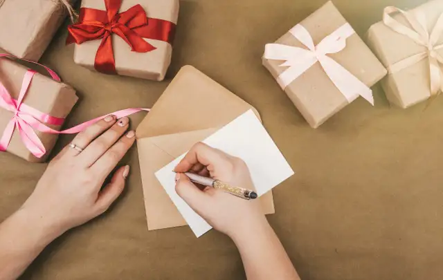 Dłonie piszą zaproszenie na kartce, obok leżą prezenty owinięte w papier i ozdobione wstążkami.