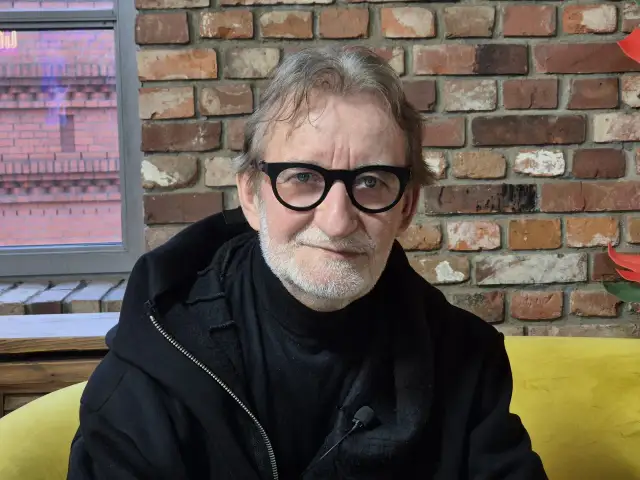 Maciej Mańkowski w okularach, z siwą brodą, w czarnej bluzie na tle ceglanej ściany.