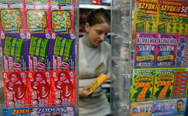 Różnorodne zdrapki loteryjne na wystawie sklepowej, w tle kobieta wybiera jedną z nich.
