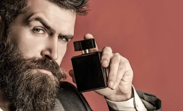 Mężczyzna z brodą trzyma czarną buteleczkę perfum na czerwonym tle.