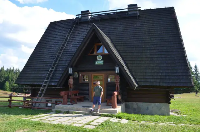 Mężczyzna stoi przed drewnianą bacówką Krawców Wierch. Dach kryty gontem, pochyła drabina.