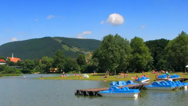 Słoneczny dzień nad jeziorem w Międzybrodziu Żywieckim. Ludzie odpoczywają na plaży, a przy brzegu cumują niebieskie rowerki wodne.